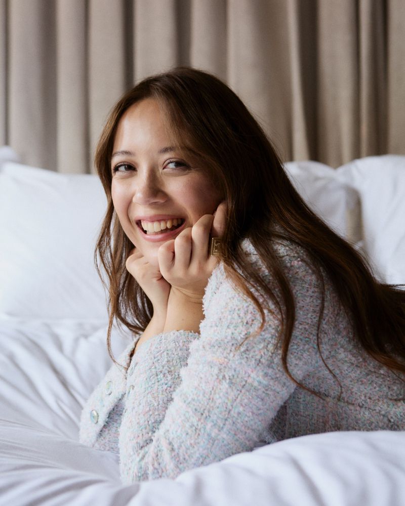 Portrait of a smiling adult woman with long brown hair, laying on white sheets wearing a textured light blue jacket indoors.