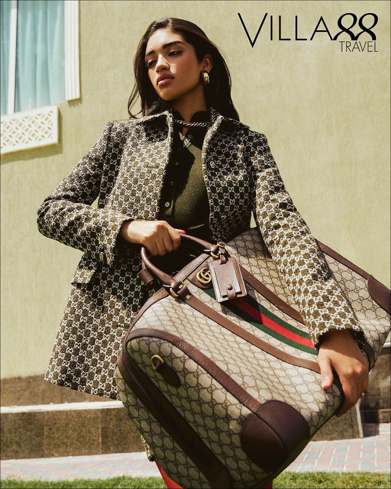 A woman carrying a Gucci travel bag. She's wearing a patterned jacket and green top, complemented by gold jewelry in an outdoor setting.
