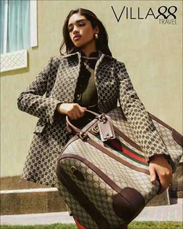 A woman carrying a Gucci travel bag. She's wearing a patterned jacket and green top, complemented by gold jewelry in an outdoor setting.