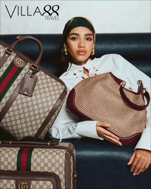 Woman poses with Gucci luggage and handbag. She is wearing a headscarf and a white blouse, photographed in a studio setting.