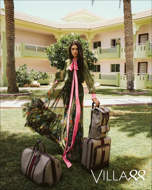 Woman in a dress with luggage in front of a building and palm trees in the background, on a sunny day.