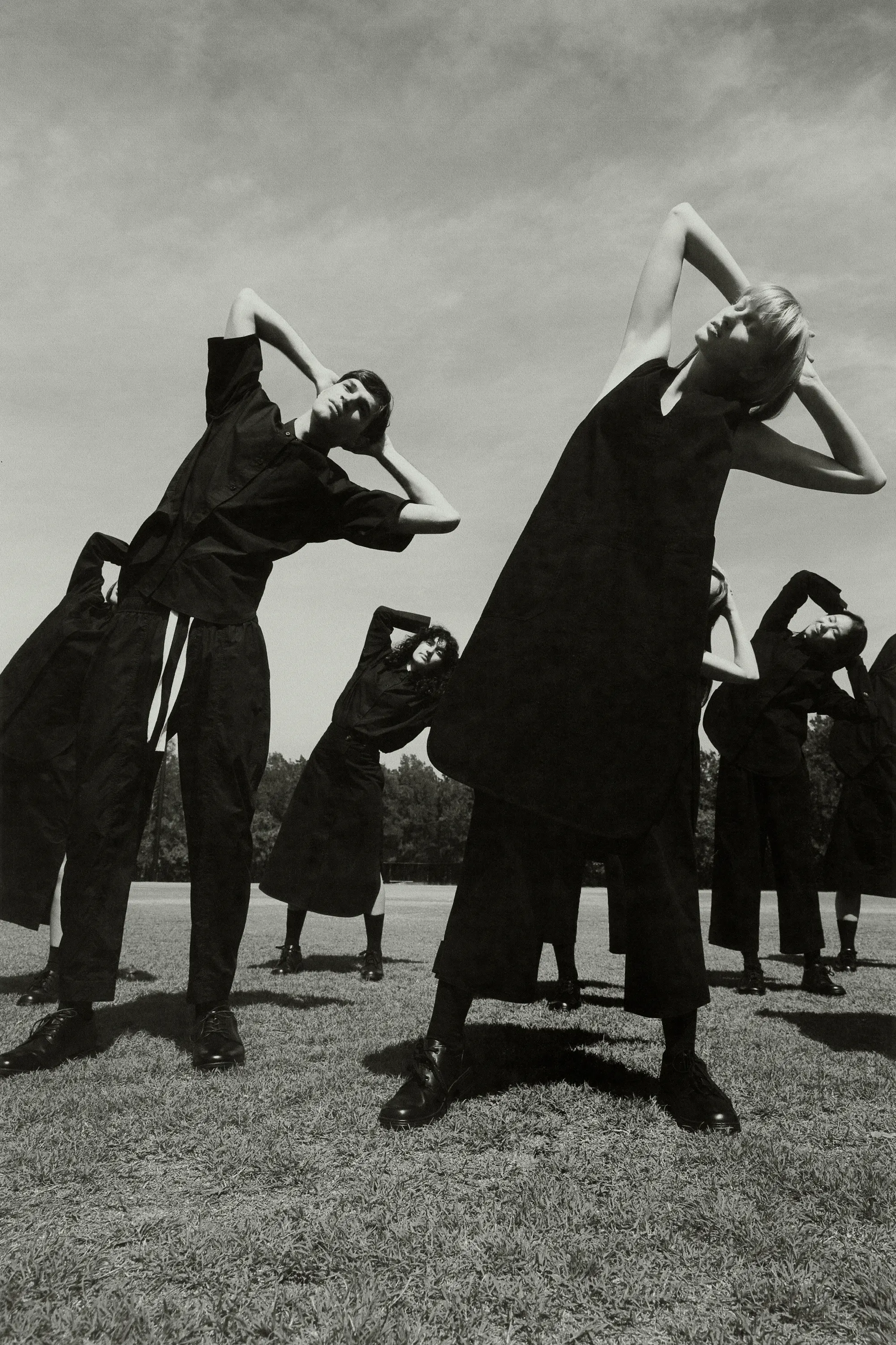 Group of models in dark clothing posing with arms raised on a grassy field under an overcast sky, photographed by Levon Baird.