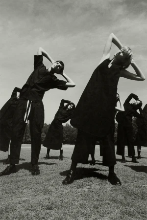 Group of models in dark clothing posing with arms raised on a grassy field under an overcast sky, photographed by Levon Baird.