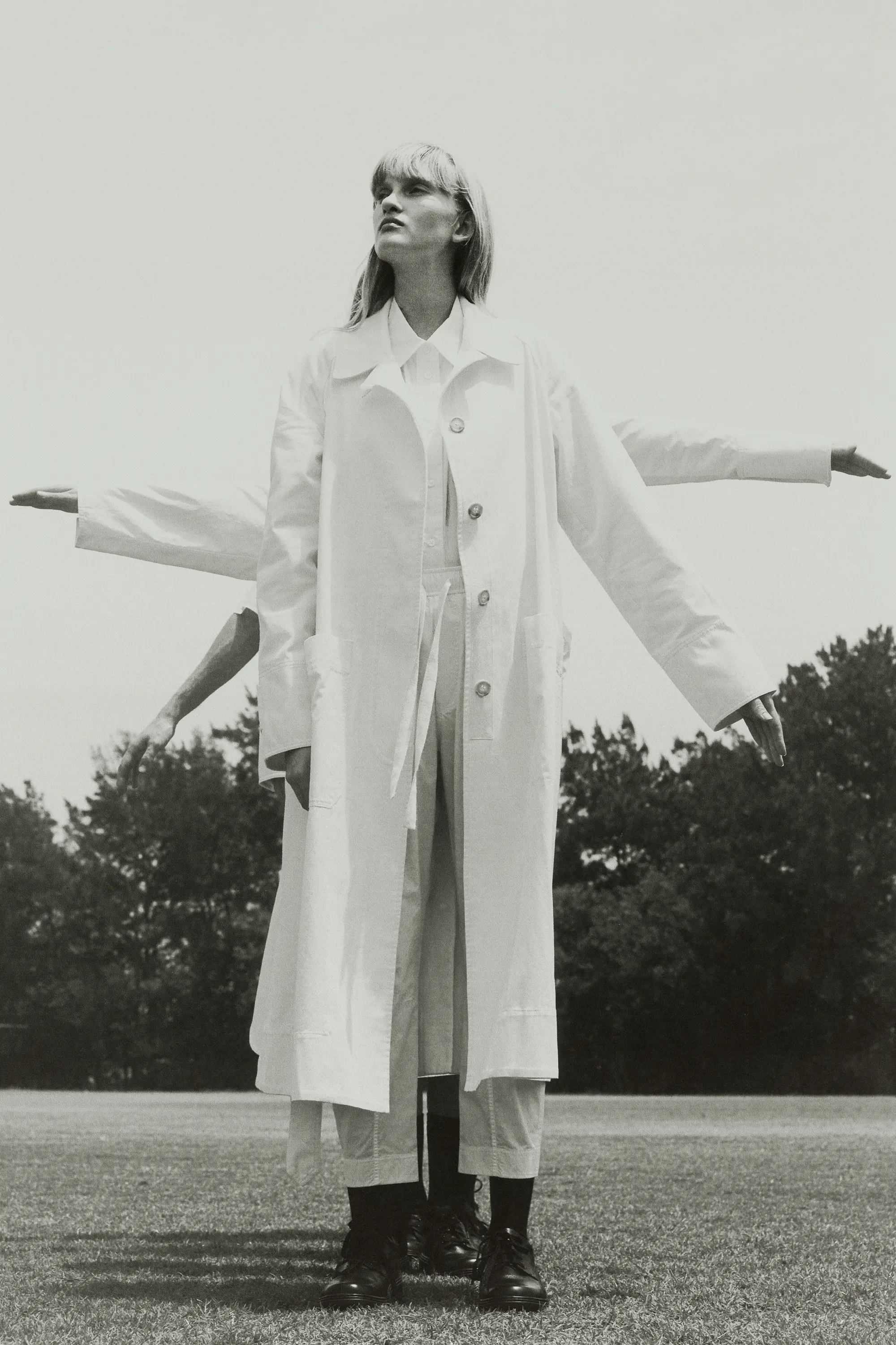 A monochrome fashion shot features a model in a white coat standing on a grassy field with duplicate arm poses against a backdrop of trees.