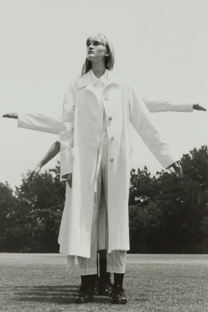 A monochrome fashion shot features a model in a white coat standing on a grassy field with duplicate arm poses against a backdrop of trees.