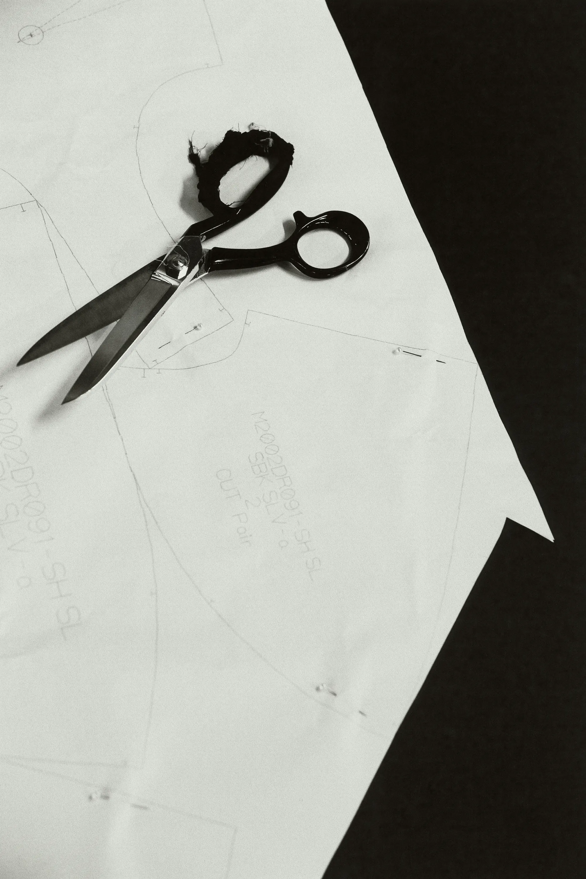 Overhead studio shot of paper sewing pattern and scissors on a black surface, emphasizing tailoring and monochrome visual texture.