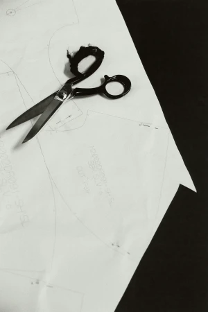 Overhead studio shot of paper sewing pattern and scissors on a black surface, emphasizing tailoring and monochrome visual texture.