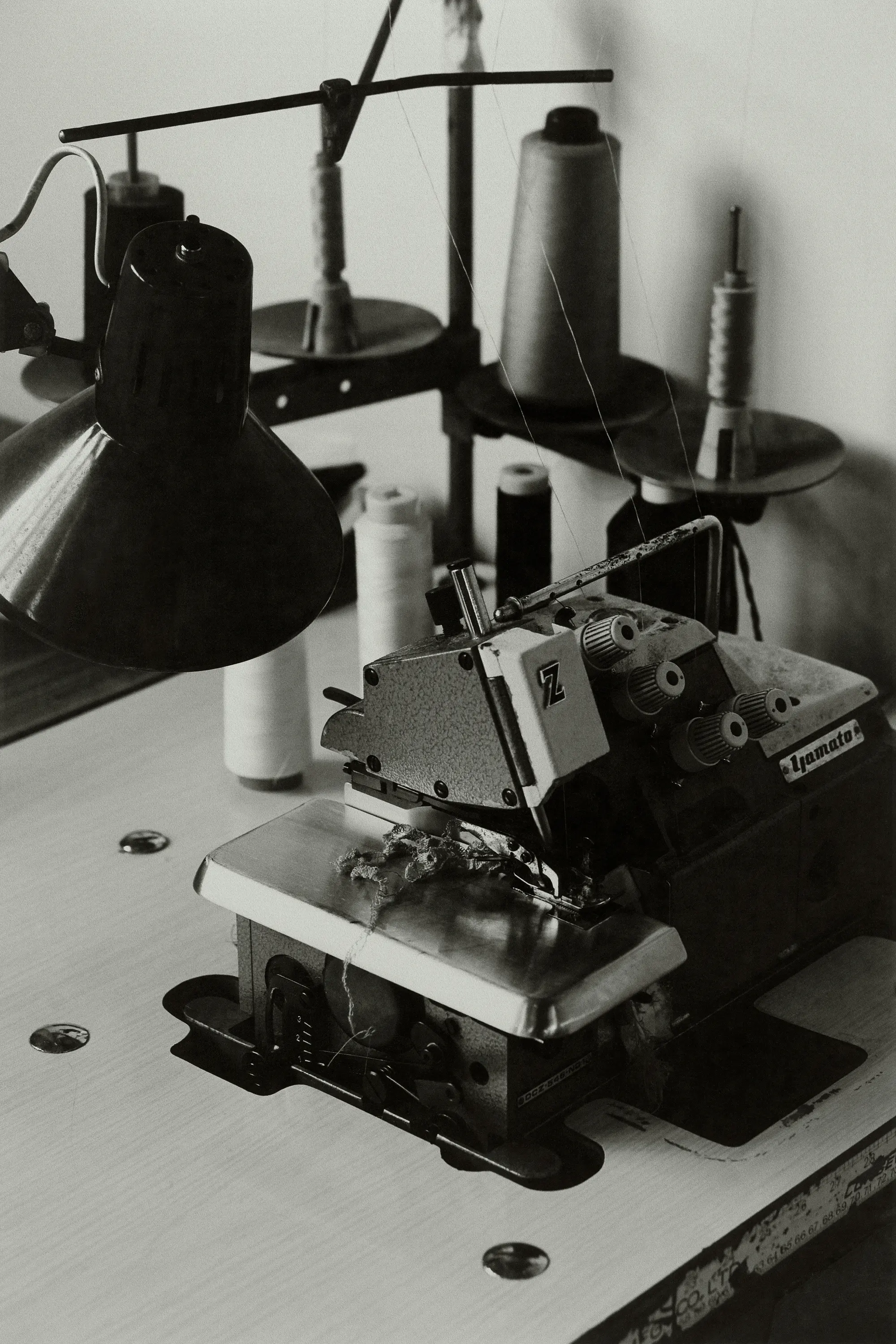 Vintage sewing machine with cones of thread and a desk lamp in a workshop, captured in black and white with shallow depth of field.