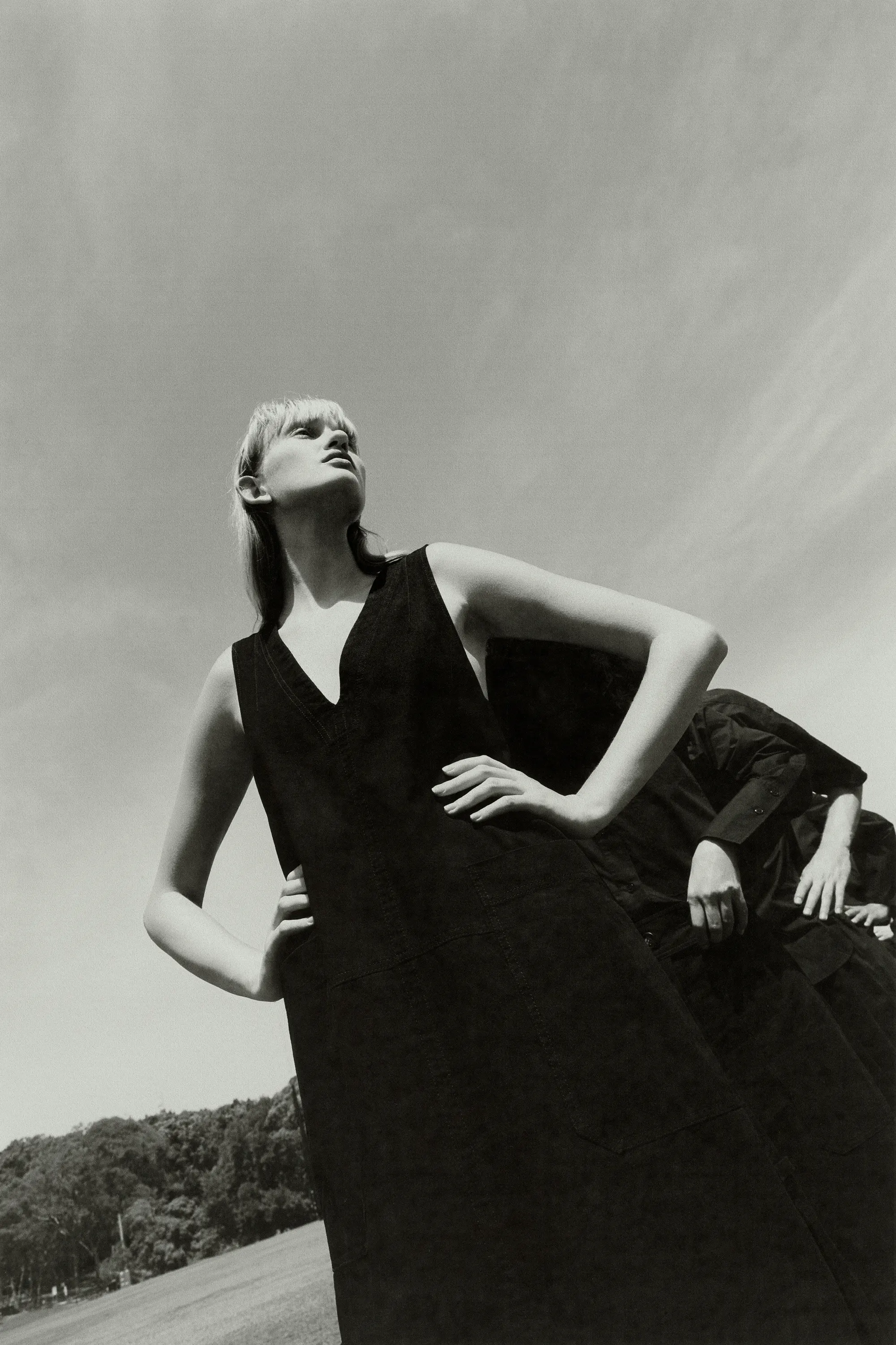 Model in dark sleeveless dress with hands on hips viewed from low angle against open sky and distant trees
