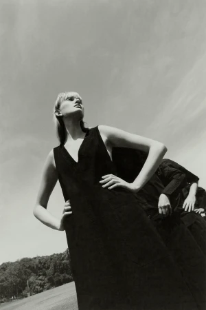 Model in dark sleeveless dress with hands on hips viewed from low angle against open sky and distant trees