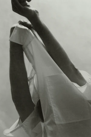 Monochrome close-up featuring a person in a white button-down shirt with arms raised against a bright sky background.