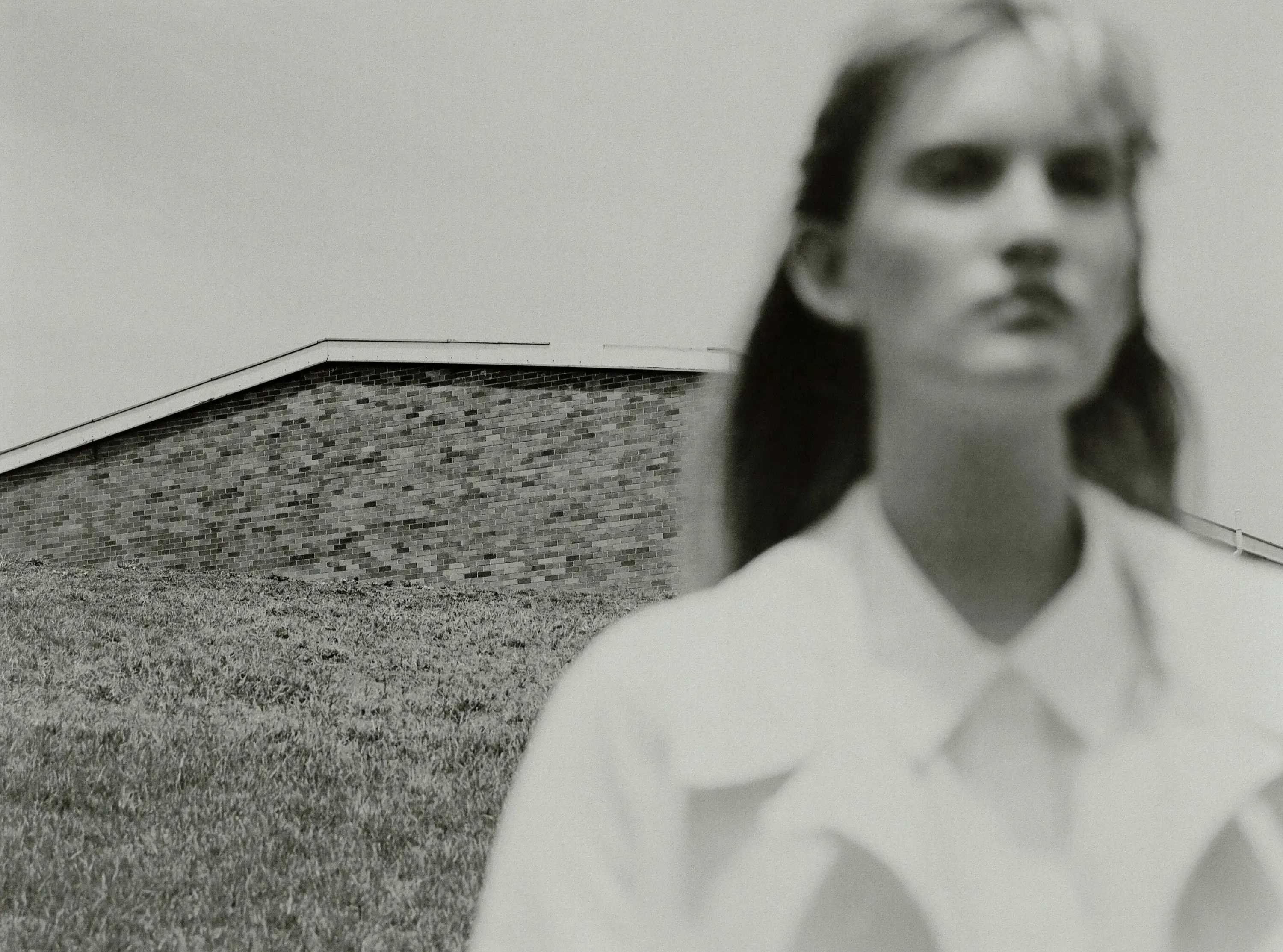 Black and white image of an adult female out of focus with a brick building and grass lawn in the background in soft light.