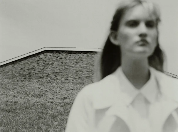 Black and white image of an adult female out of focus with a brick building and grass lawn in the background in soft light.