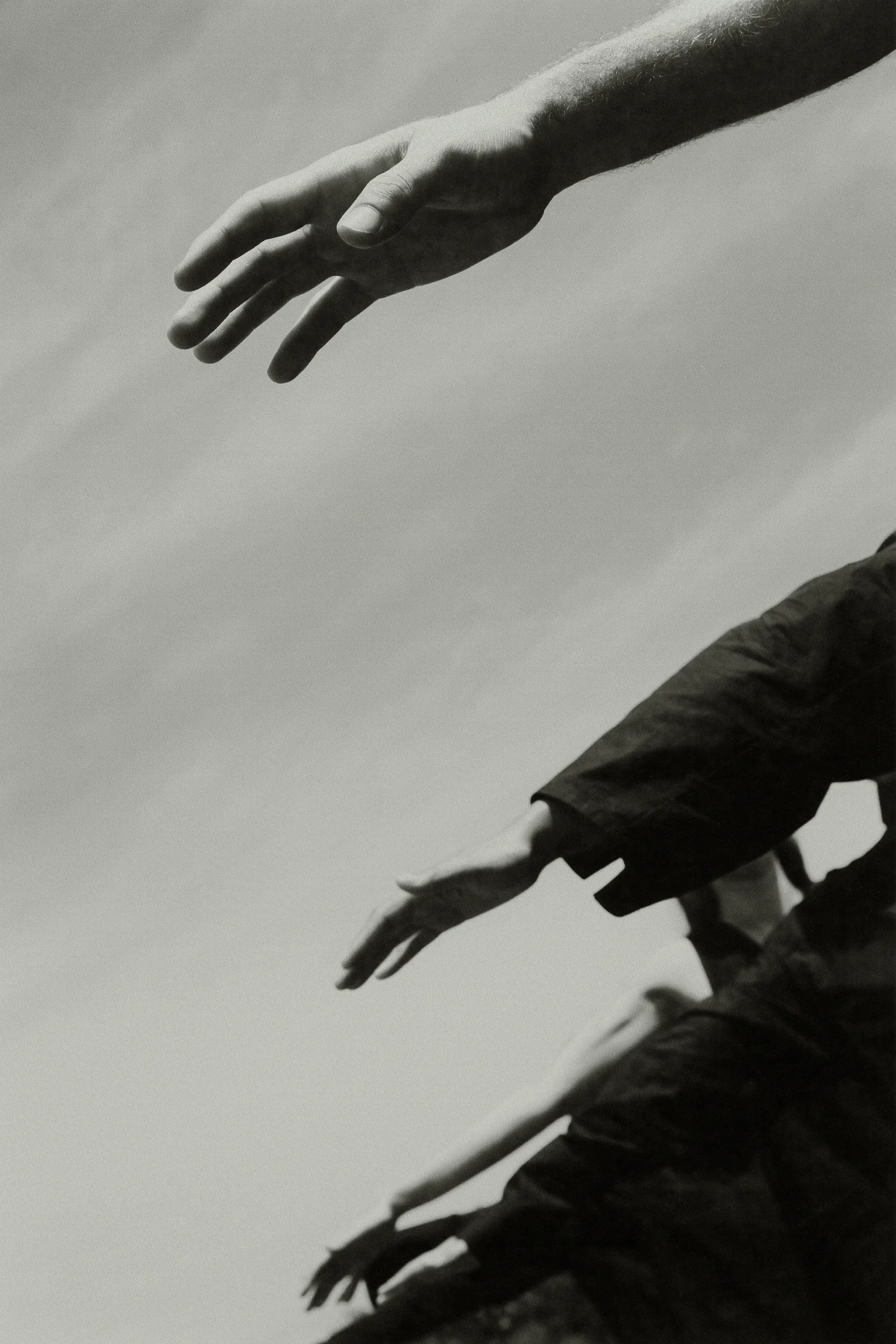 Black and white image features multiple arms and hands reaching upwards towards a bright, cloudy sky, creating a sense of ascent.