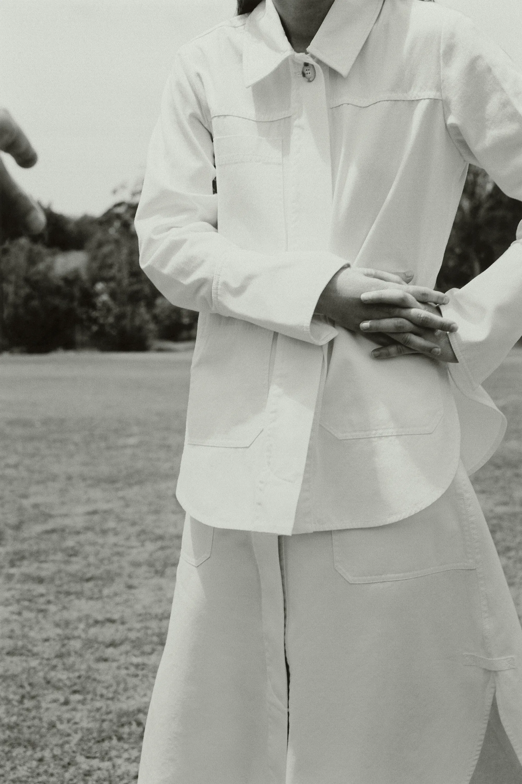Woman in a white button-down jacket and skirt standing in a grassy field, outdoors, medium shot.
