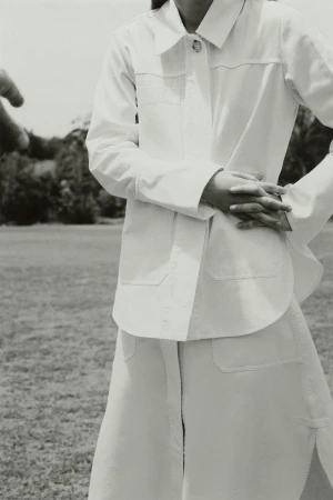 Woman in a white button-down jacket and skirt standing in a grassy field, outdoors, medium shot.