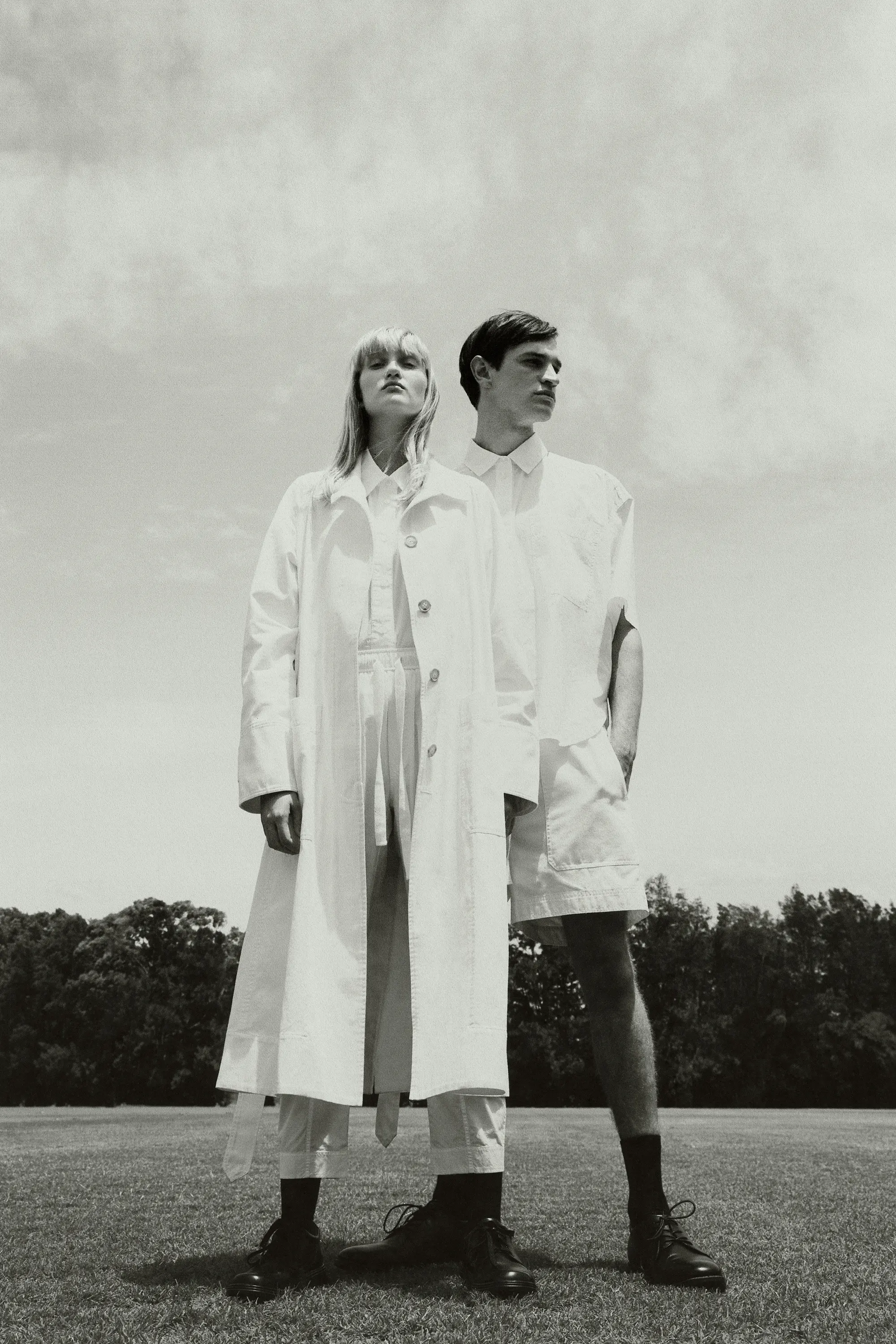 Monochromatic fashion shot of a blond woman in trench coat and a man in white separates, standing on a field in direct sunlight.