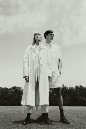 Monochromatic fashion shot of a blond woman in trench coat and a man in white separates, standing on a field in direct sunlight.