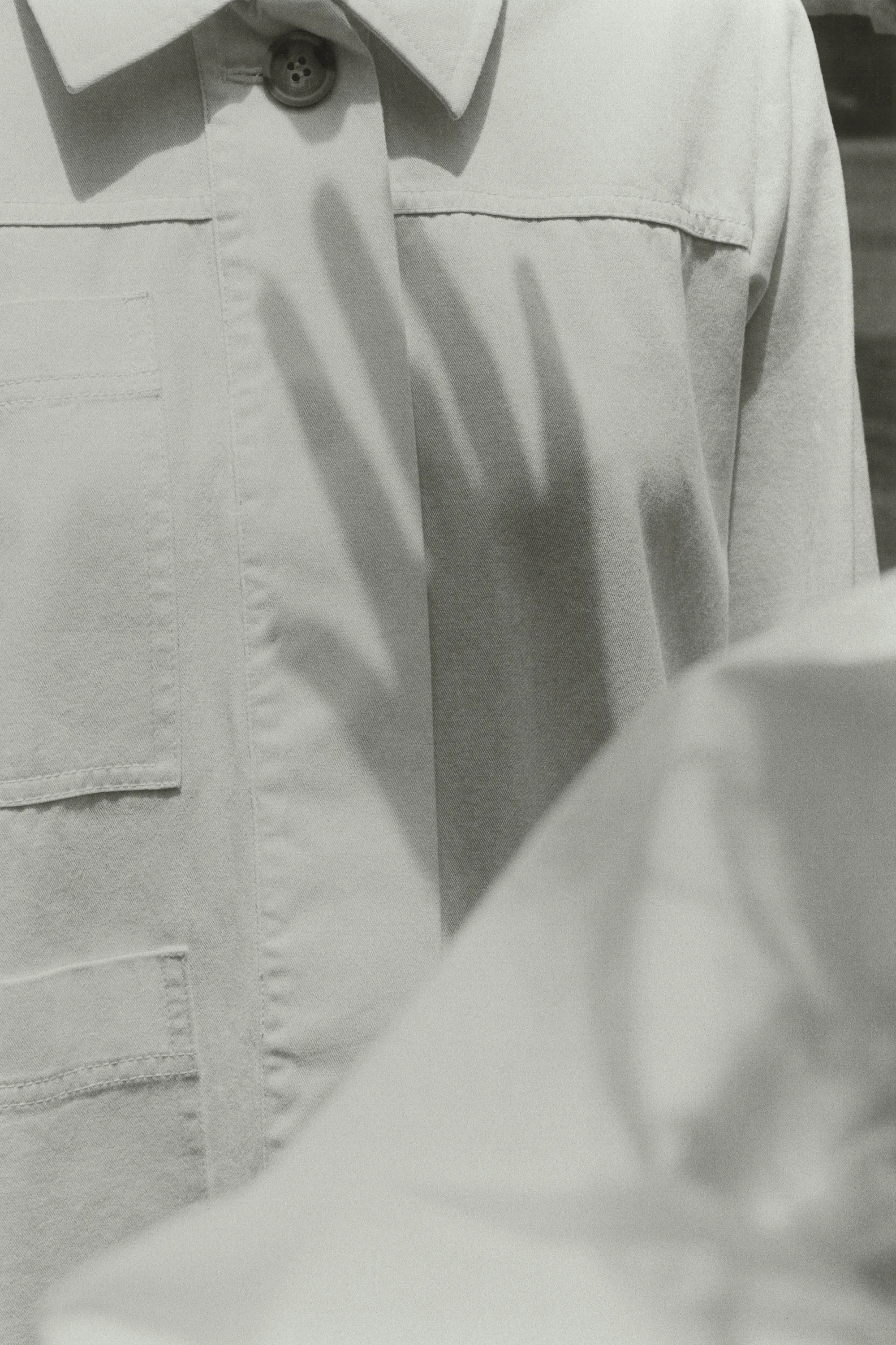 Monochrome image featuring a hand shadow cast on a tan button-down shirt with visible pockets and subtle fabric texture details.