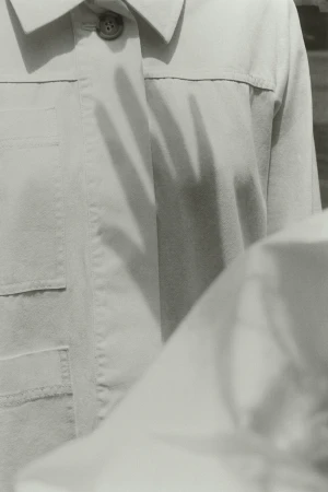 Monochrome image featuring a hand shadow cast on a tan button-down shirt with visible pockets and subtle fabric texture details.