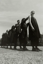 A line of models in dark clothing standing outdoors on grass under an overcast sky for Lee Mathews Workroom I.