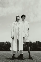 Monochrome fashion editorial featuring a standing female model in a white coat and male model on grass outdoors.
