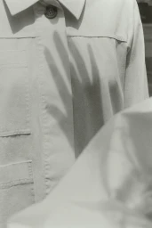 Close-up of a white utility shirt with pockets and buttons, featuring a shadow cast of a hand across the fabric.