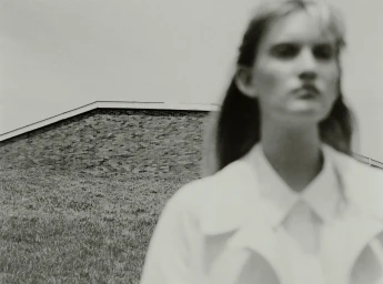 Soft-focus monochrome portrait of a model in a white shirt outdoors with a brick building and grassy hill in the background.