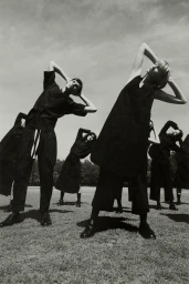 Group of models in dark clothing posing with arms raised on a grassy field under an overcast sky, photographed by Levon Baird.