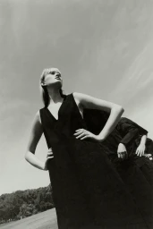 Model in dark sleeveless dress with hands on hips viewed from low angle against open sky and distant trees
