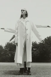 Model wearing a white long coat and trousers with outstretched arms against an outdoor grass background