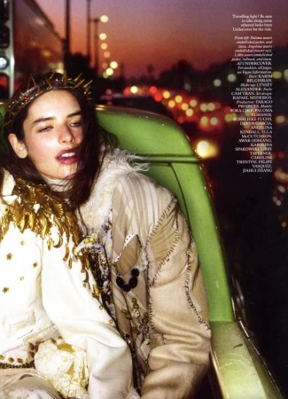 Adult female in embellished jacket and crown rides carousel at dusk with city lights in background, editorial fashion photography.
