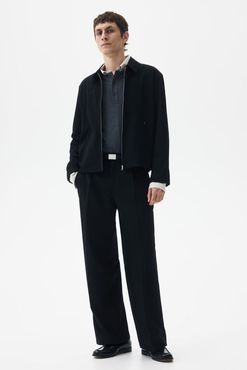 A full shot of an adult man in a black jacket and pants, posing against a white backdrop in a fashion style for a studio portrait.