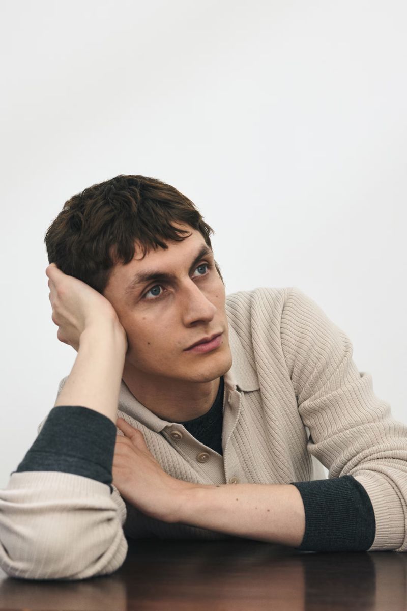 Indoor portrait of a thoughtful adult male with dark hair at a brown table, wearing a beige collared shirt against a white background.