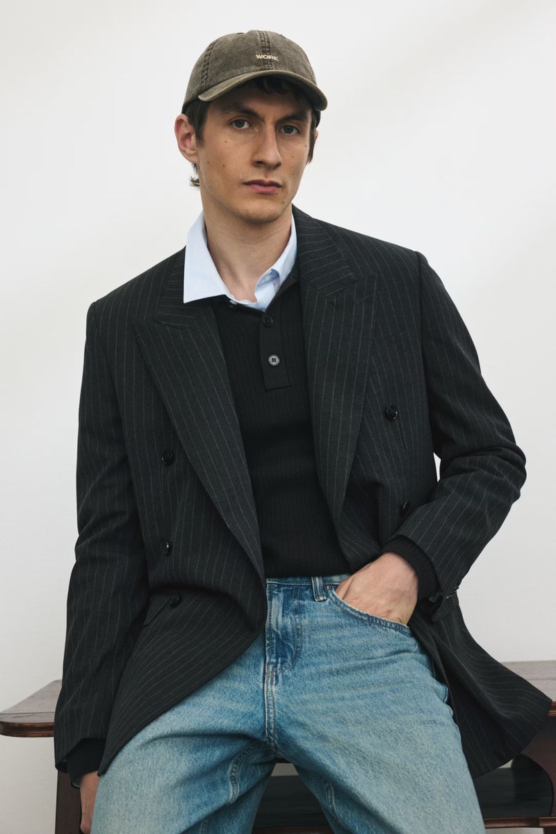 Adult male in pinstripe blazer, collared shirt, jeans, and cap poses indoors against a white wall near a wood table.