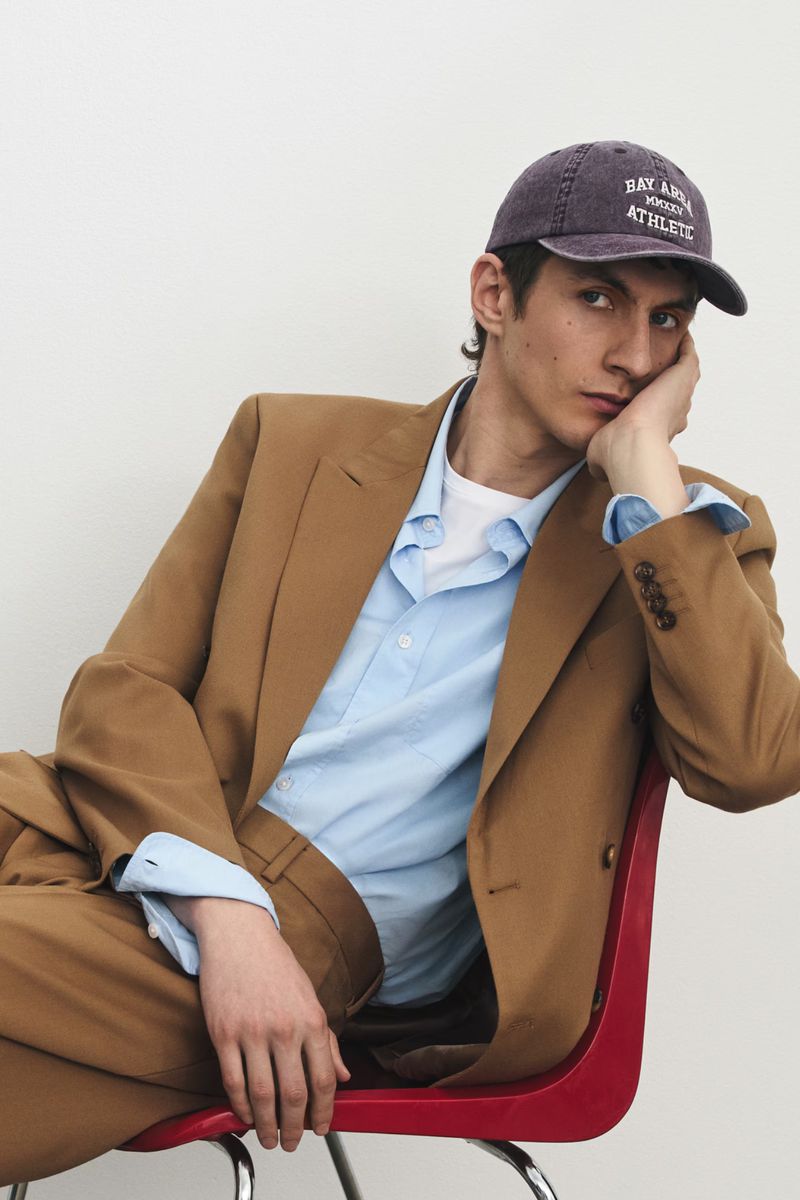 Model Henry Kitcher in a tan suit and baseball cap seated on a red chair against a white background.