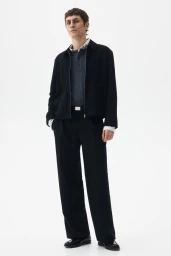 Henry Kitcher wearing a black zip-up jacket and wide-leg black trousers against a white seamless studio backdrop for H&M.