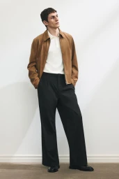 Full-length portrait of a male model wearing a brown jacket, white polo shirt, and black trousers against a neutral wall.