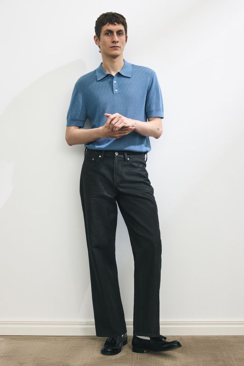 Model Henry Kitcher standing against a plain wall wearing a short-sleeve blue knit polo shirt and dark trousers.