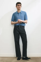 Model Henry Kitcher standing against a plain wall wearing a short-sleeve blue knit polo shirt and dark trousers.