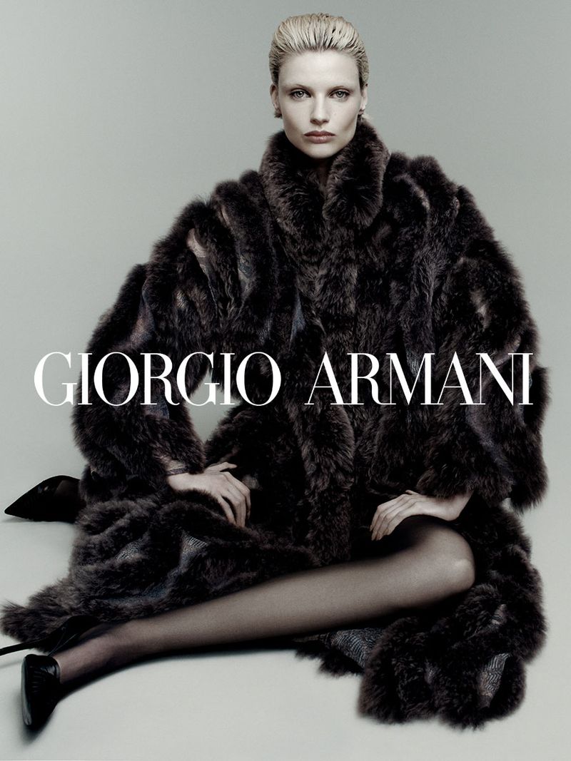 Fashion shot featuring a blonde woman wearing a large fur coat and black heels, seated against a gray background with the Giorgio Armani logo.