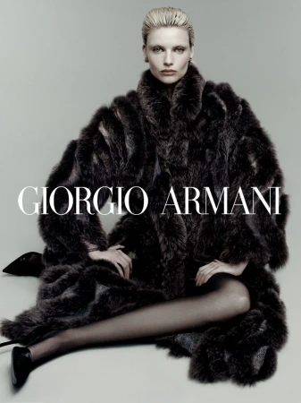 Fashion shot featuring a blonde woman wearing a large fur coat and black heels, seated against a gray background with the Giorgio Armani logo.