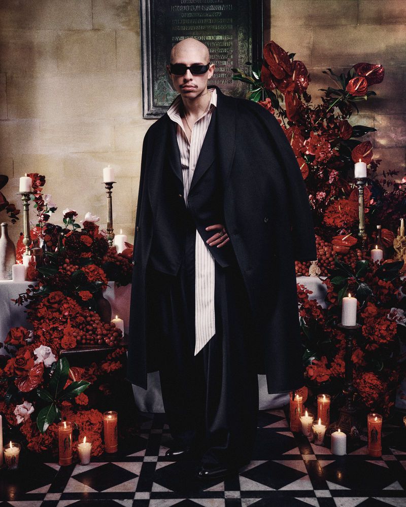 Man with shaved head and sunglasses stands in front of altar with red flowers and candles wearing a black trench coat.
