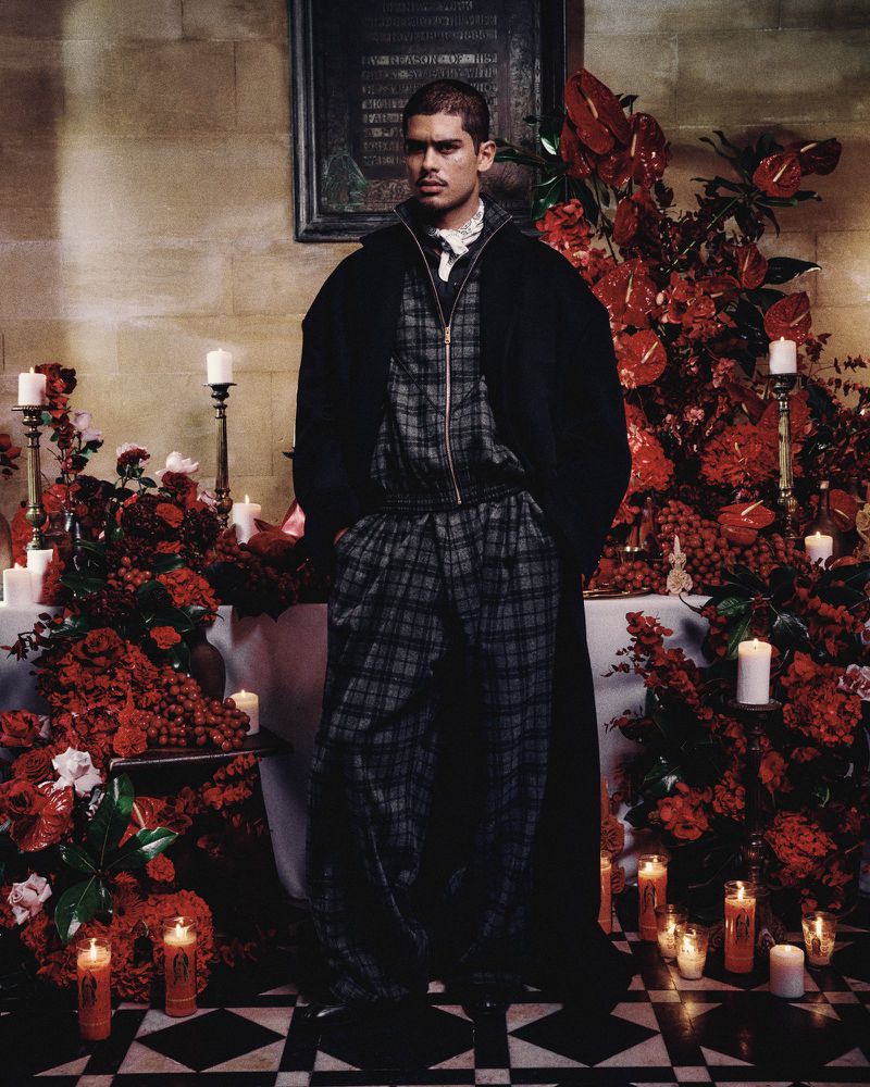 Adult male poses indoors wearing a plaid tracksuit and black coat, surrounded by red flowers and lit candles in a dimly lit room.