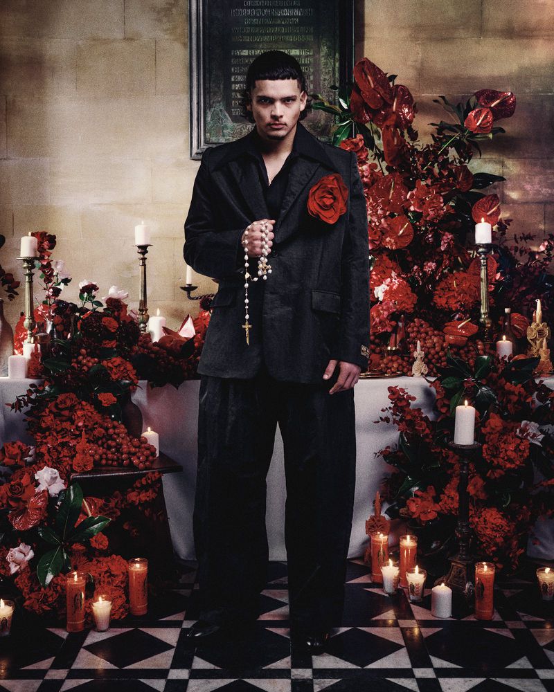 An adult male poses indoors, wearing a dark suit and holding rosary beads, set against a backdrop of red flowers and lit candles.