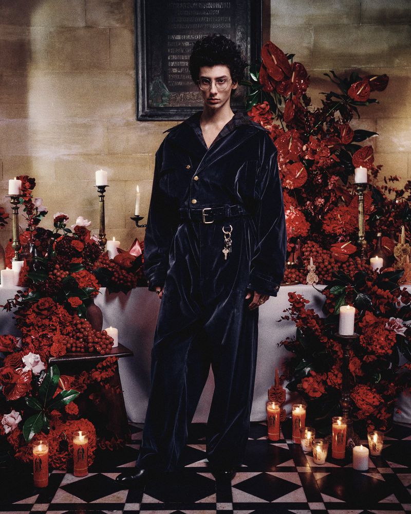 A person in a dark jumpsuit stands indoors amidst lit candles and red flowers, evoking a mysterious and dramatic ambiance.