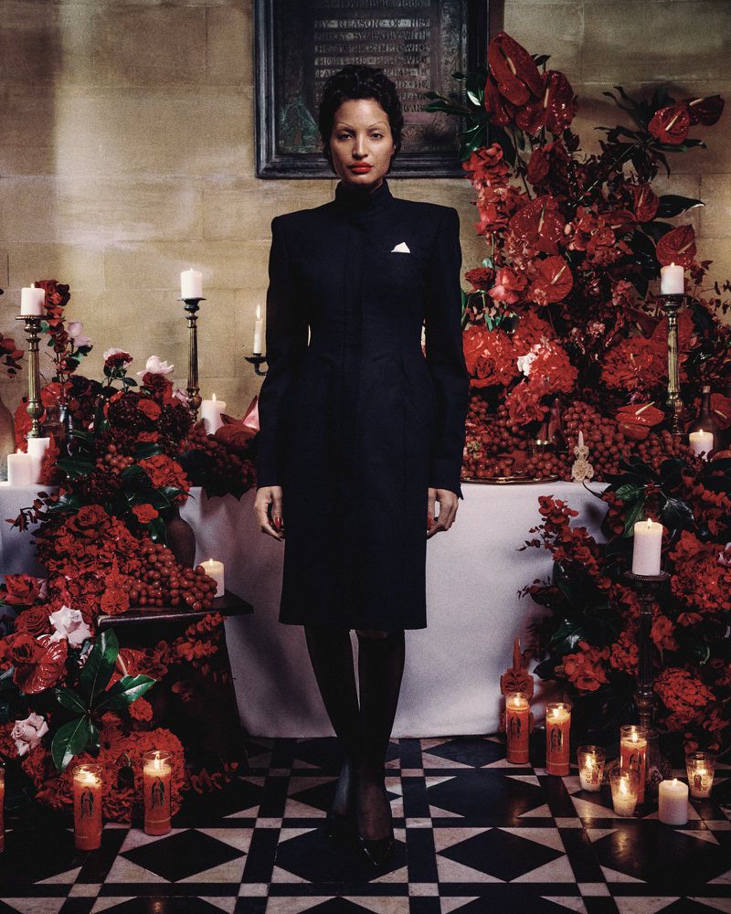 Woman in a black dress standing in a room decorated with red flowers and candles, creating a solemn and artistic ambiance.