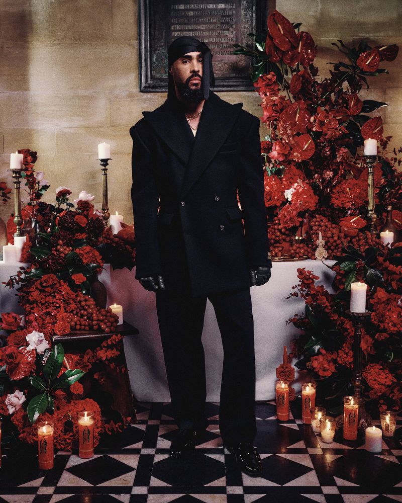 A male model in a black suit stands in front of a church altar decorated with red flowers and candles for a fashion editorial.