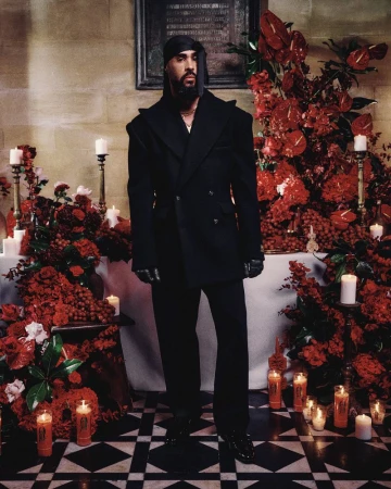 A male model in a black suit stands in front of a church altar decorated with red flowers and candles for a fashion editorial.
