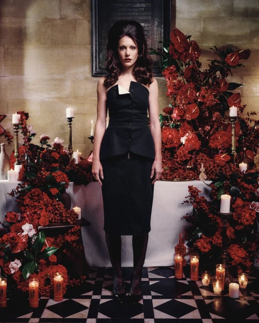 A woman in a strapless black dress stands amidst a display of red flowers and candles in a room with a black-and-white tiled floor.